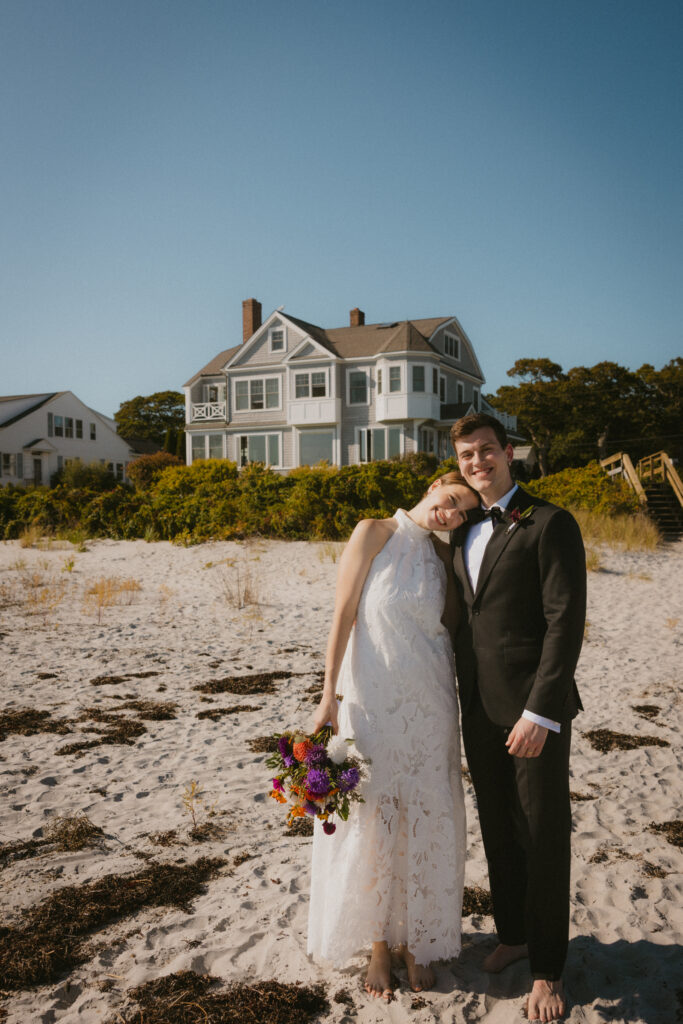 Bride and groom smiling at the camera on the beach with a classic New England beach house in the background