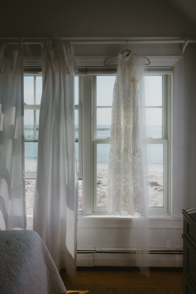 Wedding dress hanging up in front of a window with the backdrop of the  beach