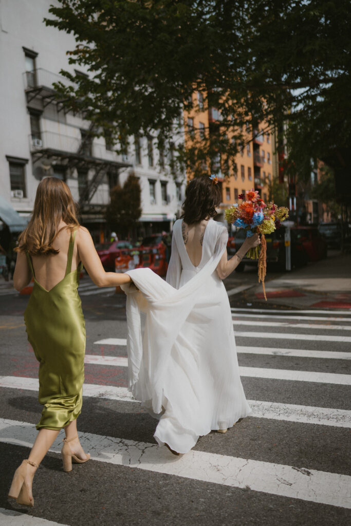 Bride walking across the street in NYC with her maid of honor holding the back of her dress