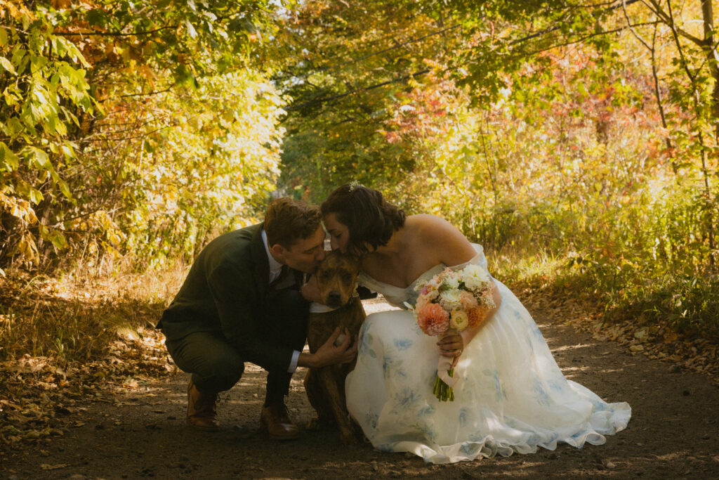 Bride and groom kissing their dog with fall foliage in the background
