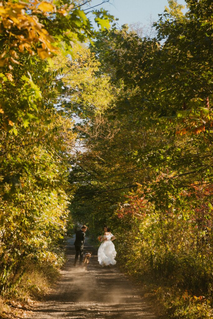 Bride and groom running down a trail with their dog surrounded by fall foliage in new england