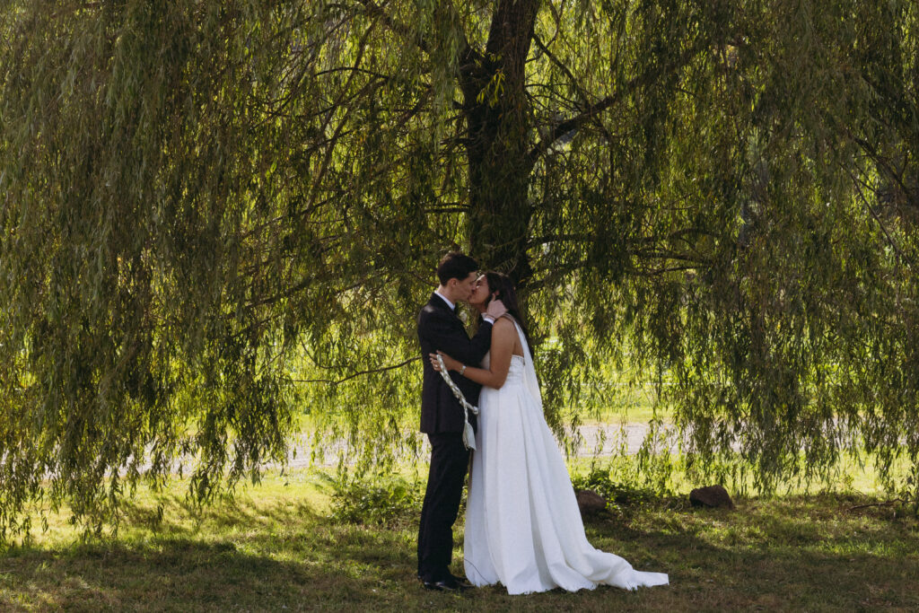 Bride and groom sharing their first kiss as a married couple in front of a willow tree