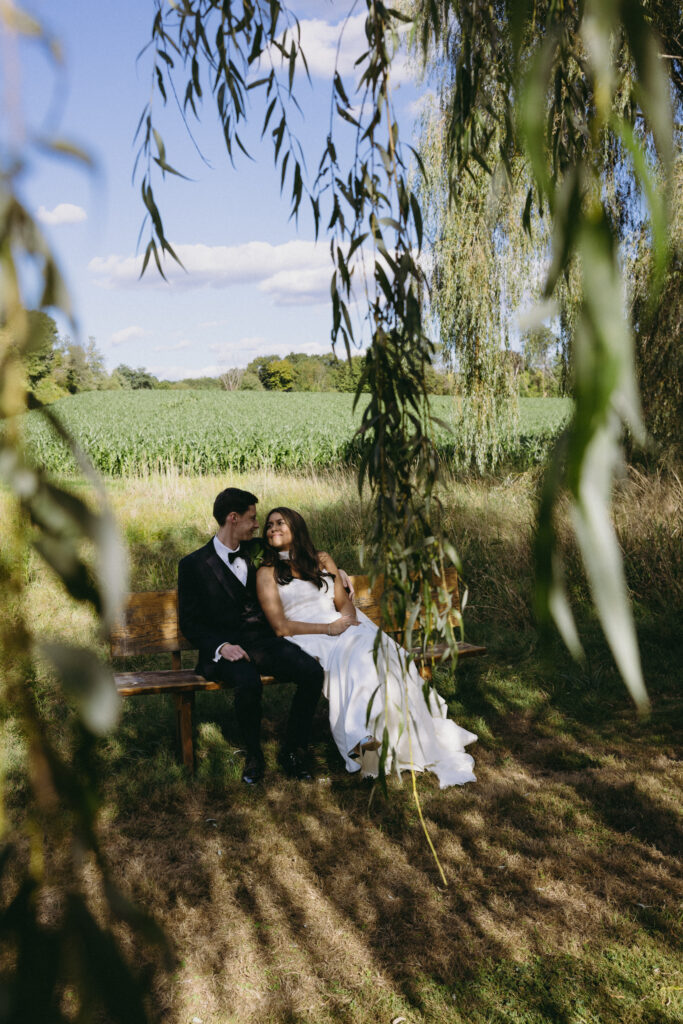 Bride and groom sitting on a bench together with willow tree leaves hanging in the foreground