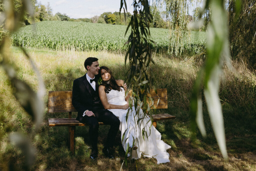 Bride and groom sitting on a bench together with willow tree leaves hanging in the foreground