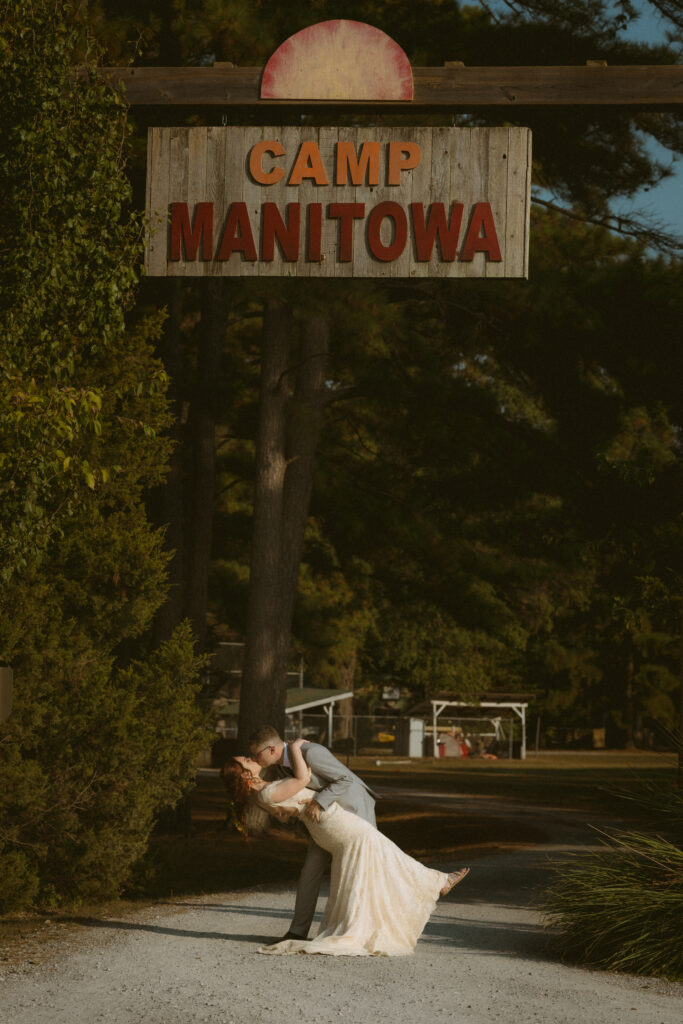 Bride and groom dip kissing in front of the camp sign at a summer camp wedding 