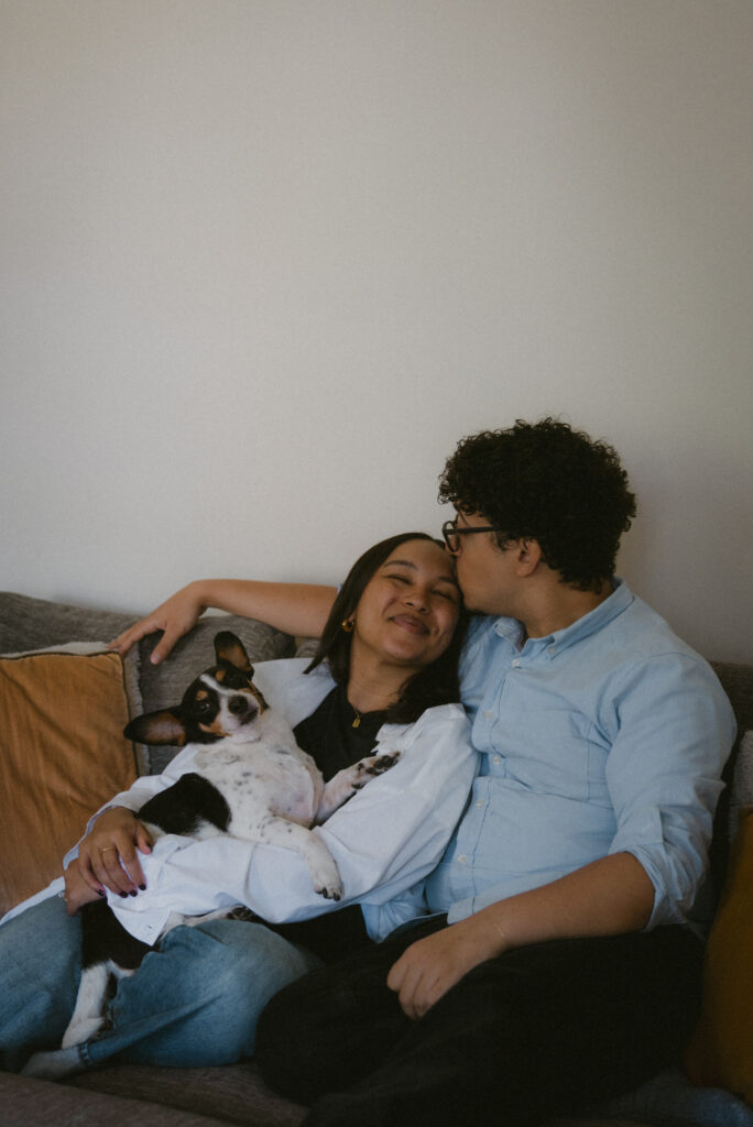 Engaged couple sitting on their living room couch holding their dog 