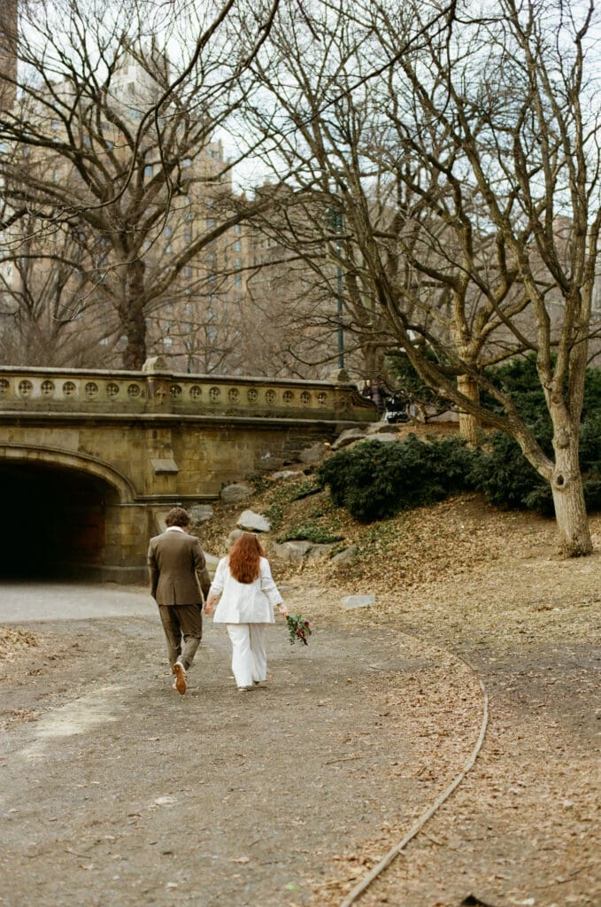 Bride and groom walking through Central Park with a bridge in the background