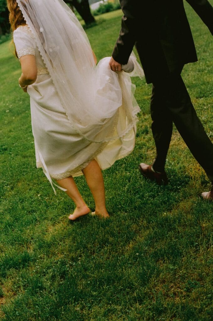 Bride and groom walking on the grass with the groom holding the brides dress and veil 