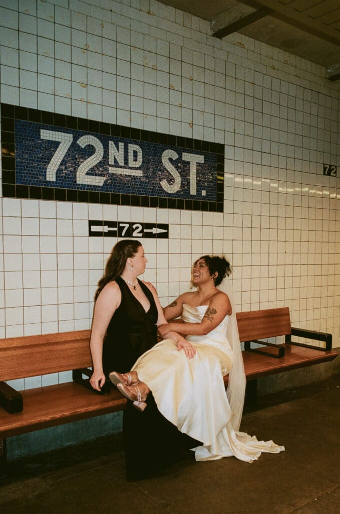 Bride resting her legs on the other bride's lap as they sit on a bench in a subway station