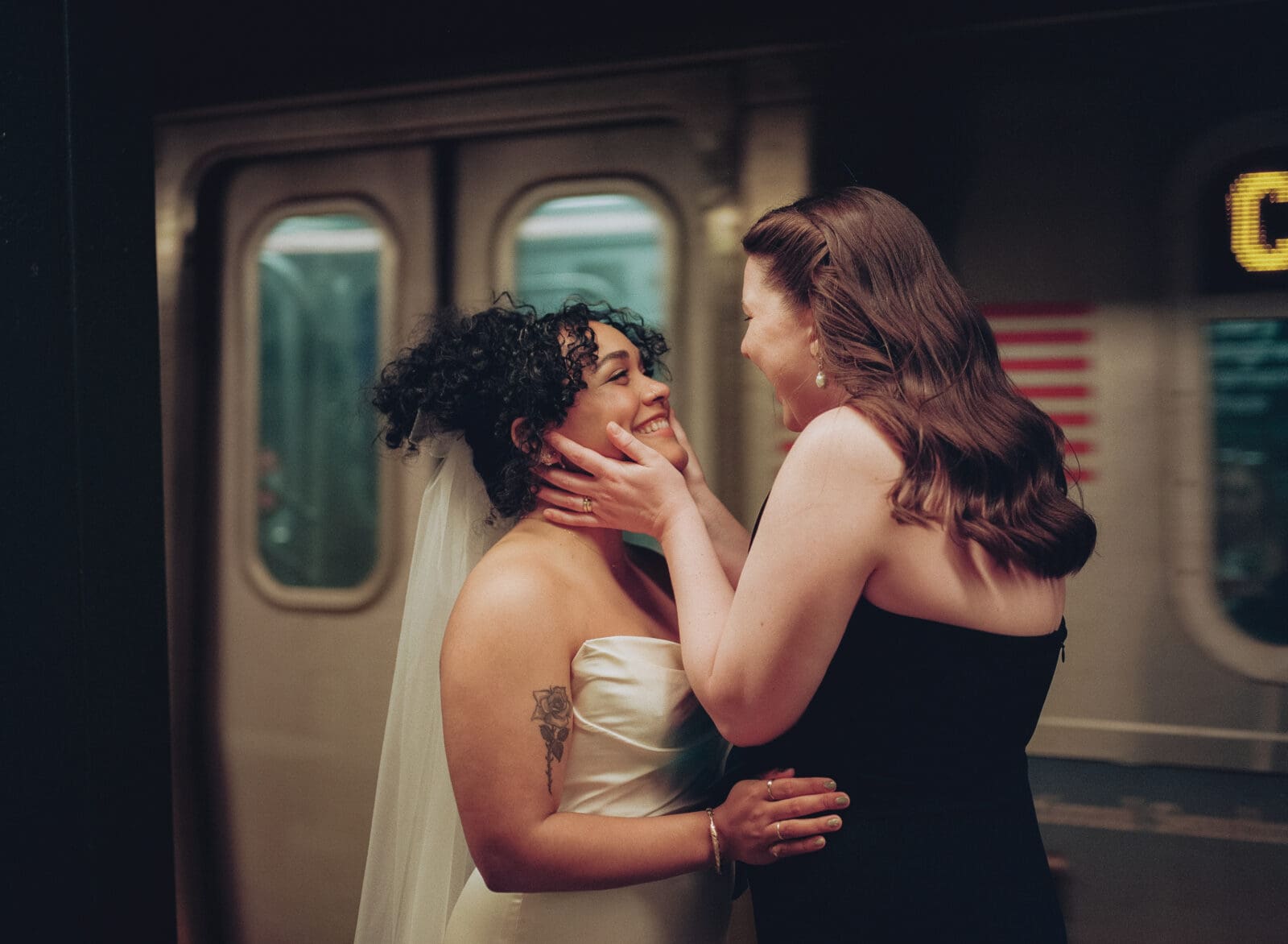 Brides smiling at each other in front of a train in the subway station with one bride holding the other's face