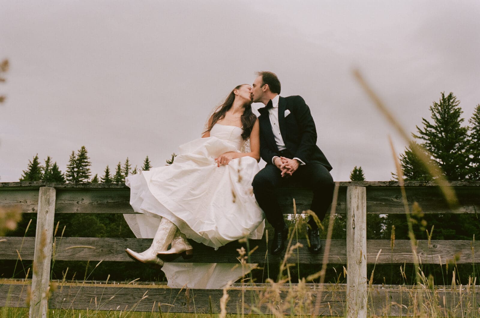 Bride and groom kissing on top of a fence in a field