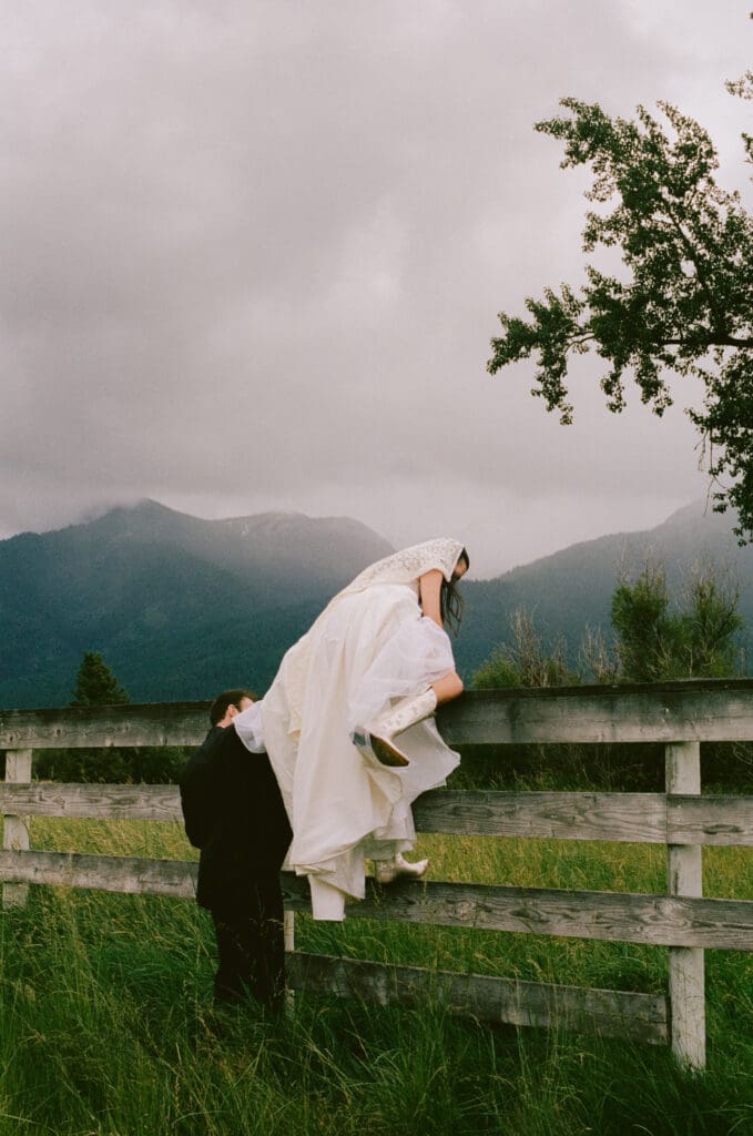 Bride climbing a fence in a field with mountains in the background
